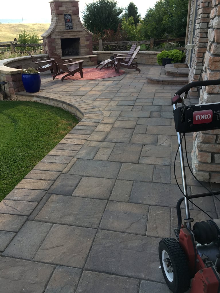 Red and black Toro lawn mower on a stone patio with outdoor fireplace, seating, and manicured lawn visible