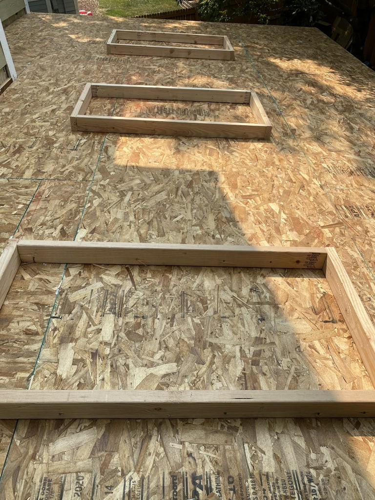 Three wooden raised garden bed frames arranged diagonally on a plywood ground in a backyard