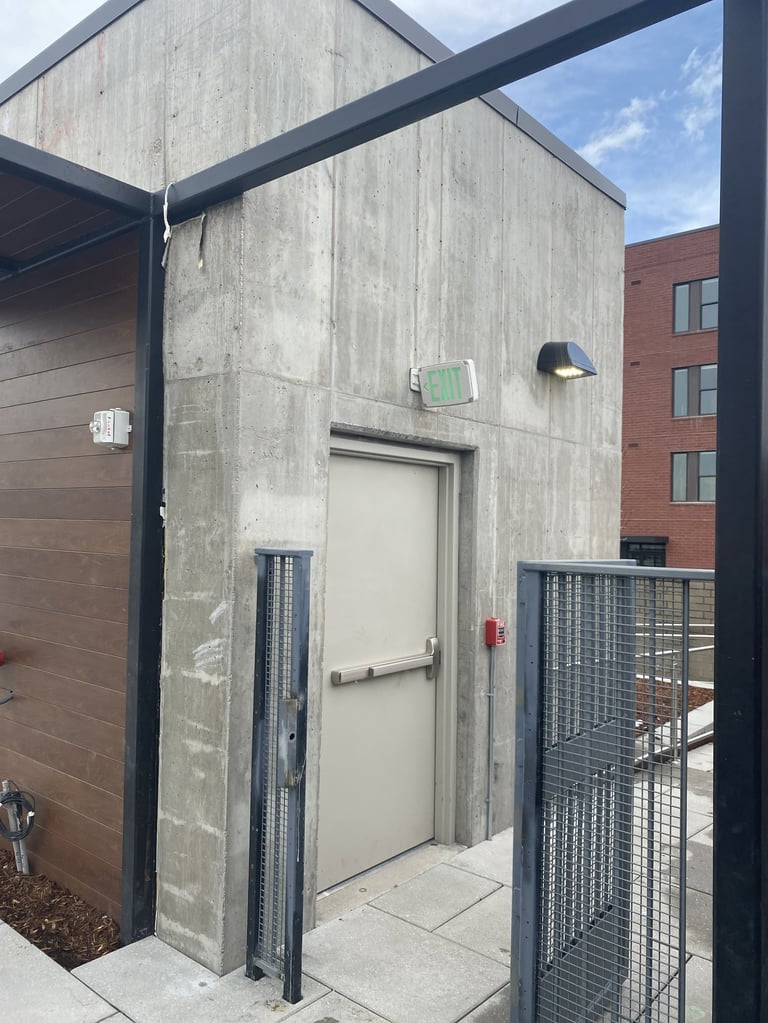 Concrete utility building with white door and metal frame on urban rooftop next to brick building
