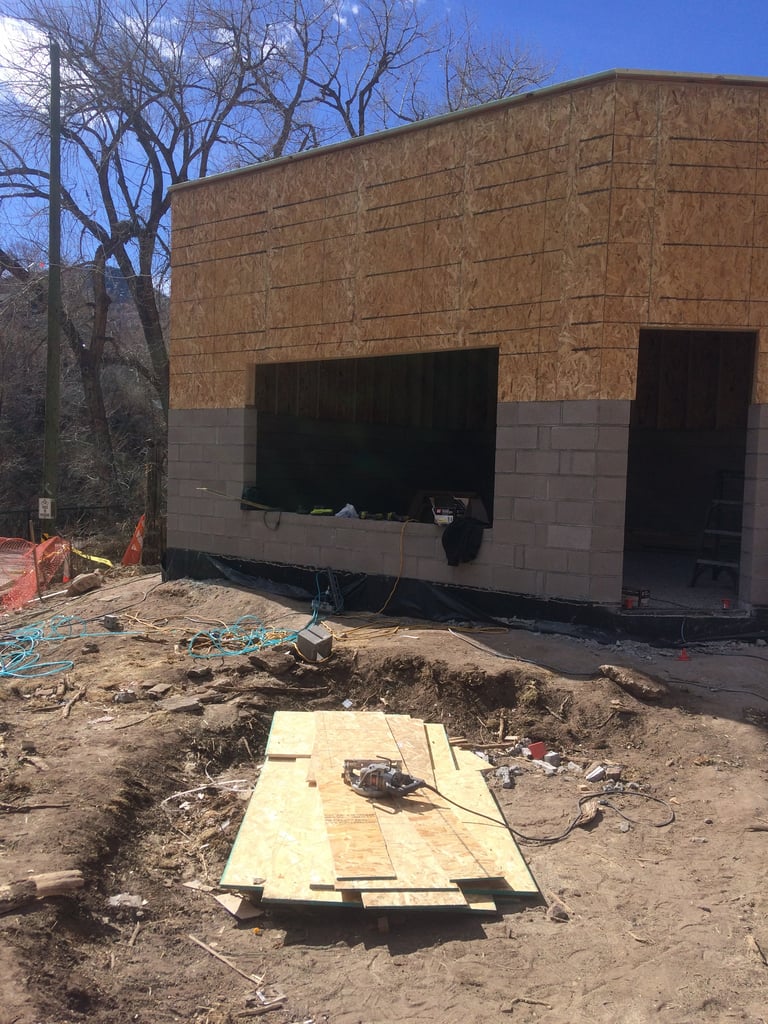 Construction site showing brick building under renovation with exposed foundation, plywood laid on ground, and bare tree in background on clear day