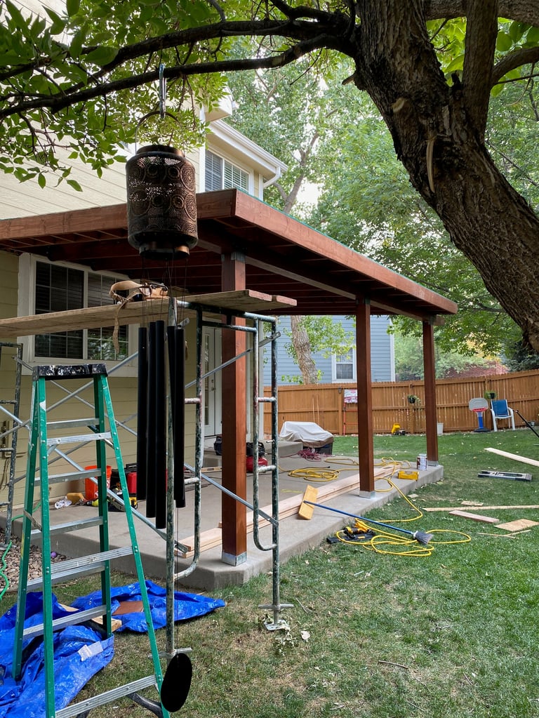 Backyard patio under construction with wooden pergola, large tree overhead, ladder, and tools scattered in yard