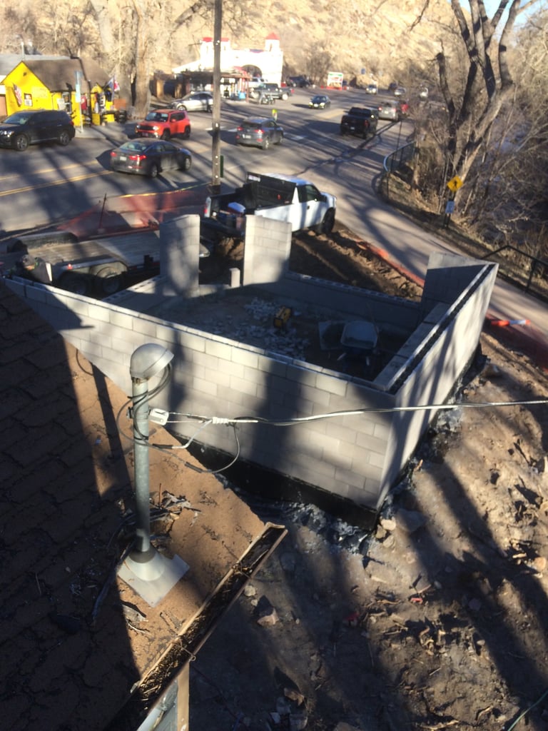 Construction site with metal dumpsters and equipment, trees, and vehicles in background on sunny day