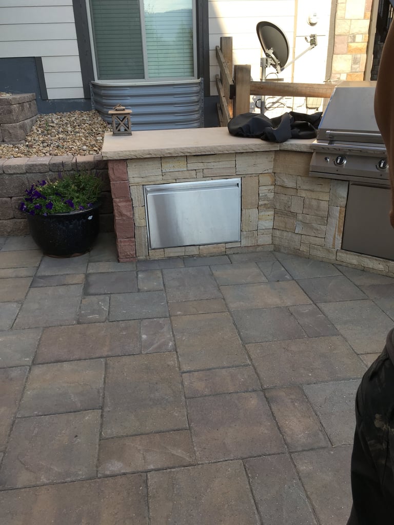 Outdoor patio with stone countertop, built-in grill, concrete pavers, and potted flowers along a residential home