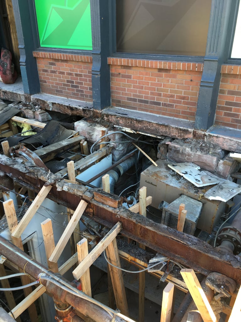 Construction site showing excavated storefront foundation with wooden support beams, metal framework, and brick building base