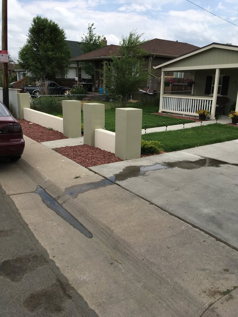 Residential street with white concrete bollards in front yard, two single-story houses with porches, green lawn, red mulch beds, and parked car