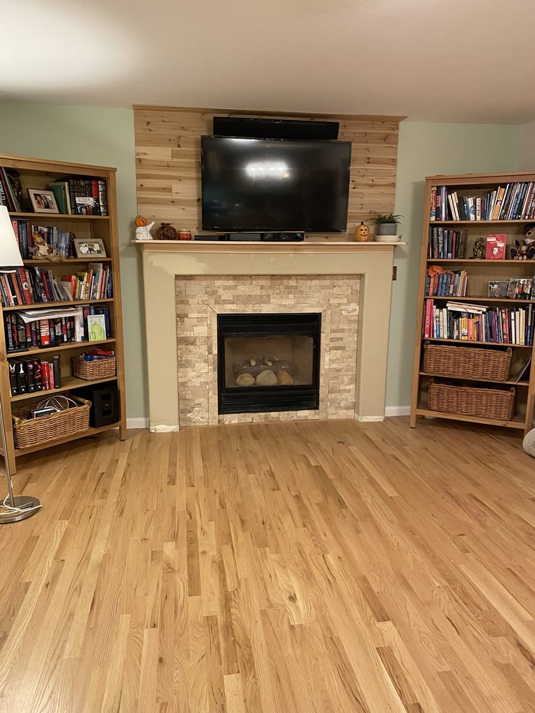 Living room with wooden fireplace, mounted TV, and bookshelves on either side above hardwood flooring