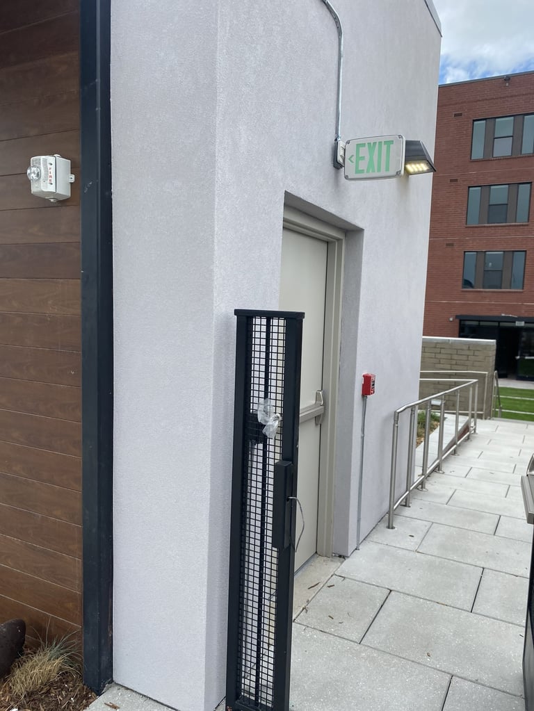 White building exterior with green EXIT sign, gray door, black gate, and brick building visible in background