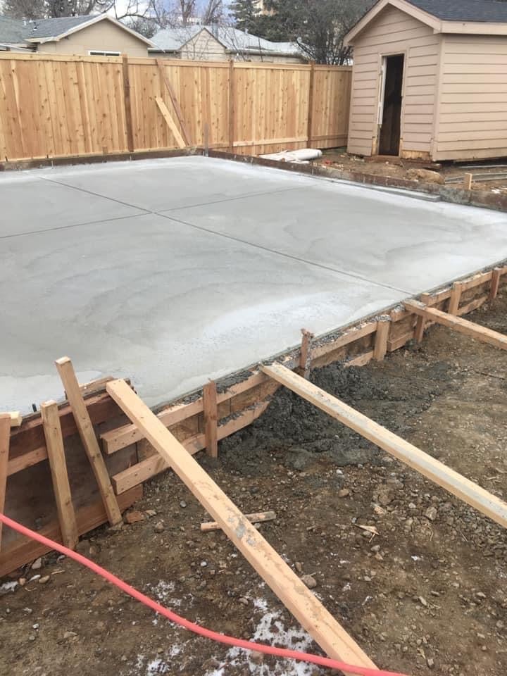 Backyard patio under construction with freshly poured concrete slab and wooden framing, wooden fence and shed in background