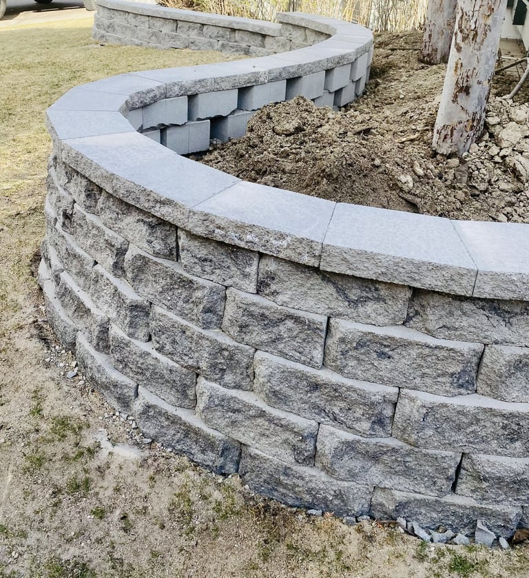 Curved retaining wall made of gray stacked stone blocks with coping cap, under construction in landscaped yard