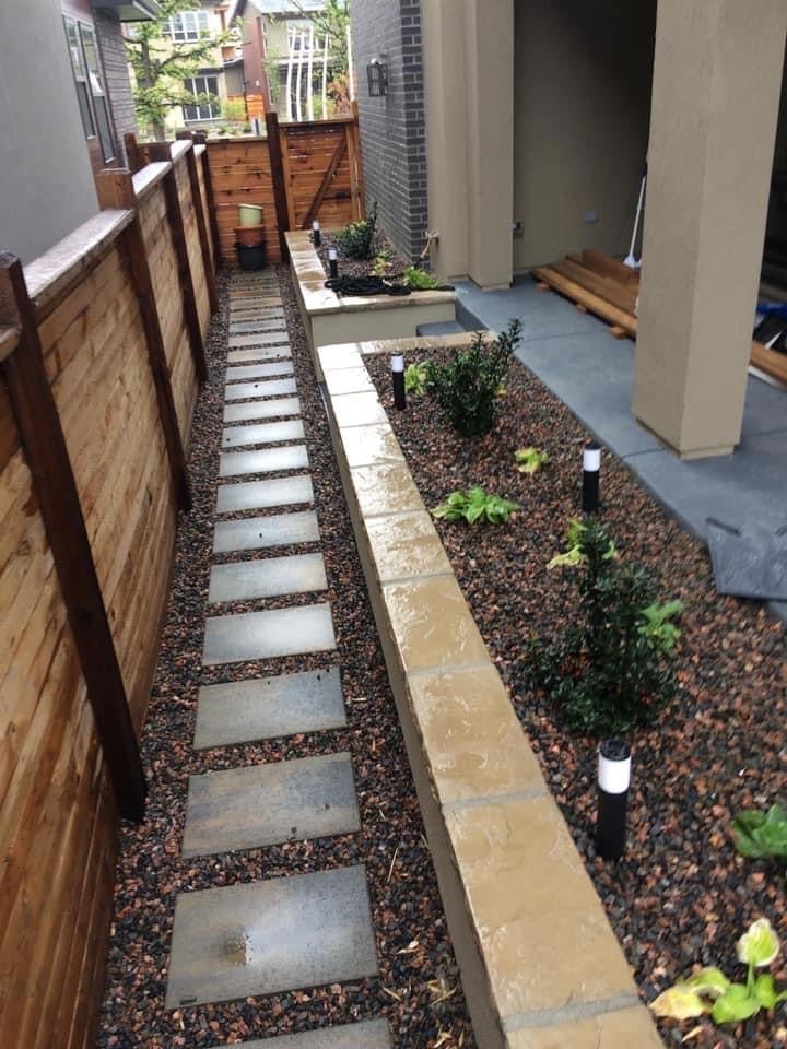 Narrow residential side yard with stepping stone pathway, gravel landscaping, young plants, wooden fence, and brick building wall