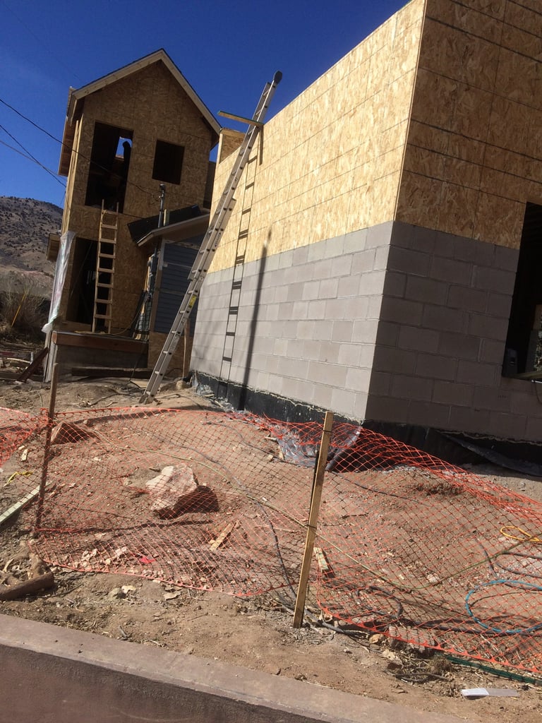 Residential building under construction with wooden framing, partial concrete block walls, and metal ladders on sandy red soil site